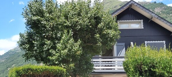 Un chalet en bois sombre doté d’un balcon, entouré d’une végétation luxuriante et adossé à un paysage montagneux.