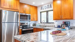 Una cocina con armarios de madera, electrodomésticos de acero inoxidable, encimera de granito y un ventanal con la palabra "ALOHA".