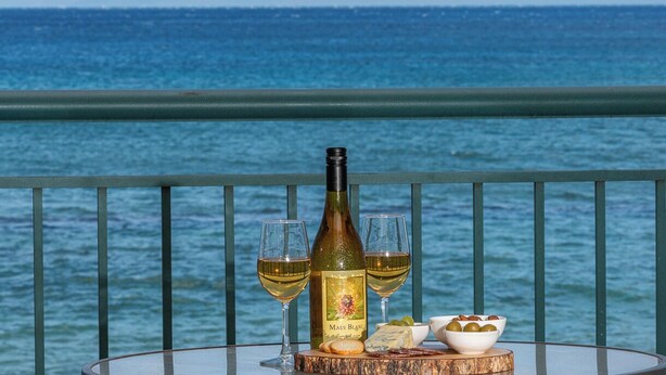 Una mesa con una botella de vino, dos copas, queso, aceitunas y galletas en un balcón con vistas al mar.