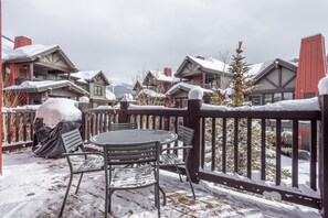 Ein mit Schnee bedeckter Balkon mit einem runden Tisch und Stühlen, umgeben von Holzwerteln und verschneiten Gebäuden im Hintergrund.
