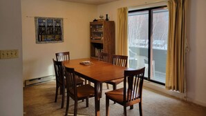 A dining room with a wooden table and chairs, a bookshelf, a quilted artwork, and a window with curtains.