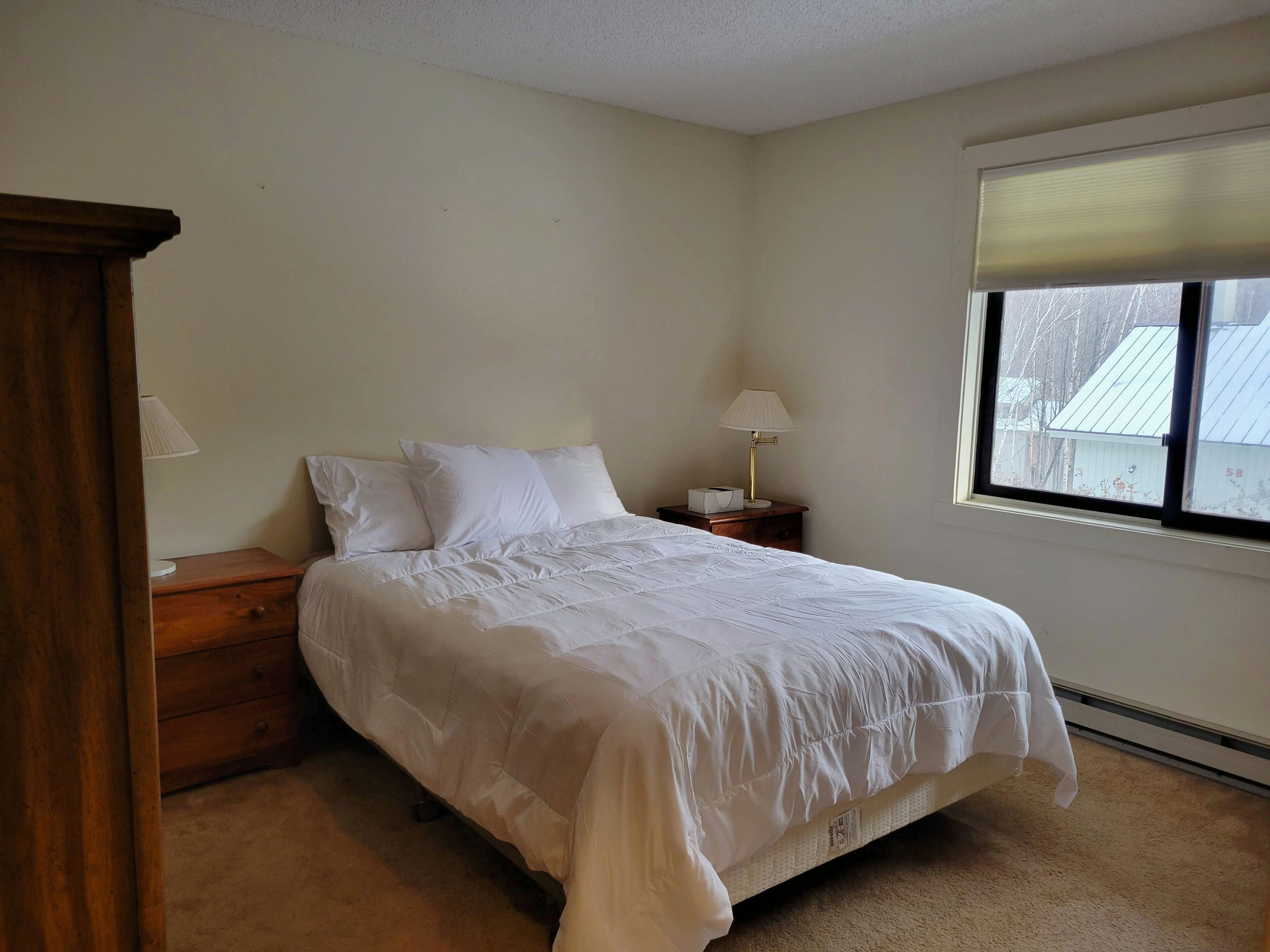 Quarto com cama, mesas de cabeceira de madeira, abajur, janela com persianas e vista para o exterior.