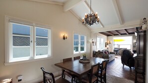 A dining area with a wooden table and chairs, large windows, a chandelier, and a view into the living room. - 1205O Spanish Beach Bungalow Retreat (Oxnard)
