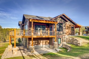 A two-story house with a balcony, stone foundation, and a view of a forested area.