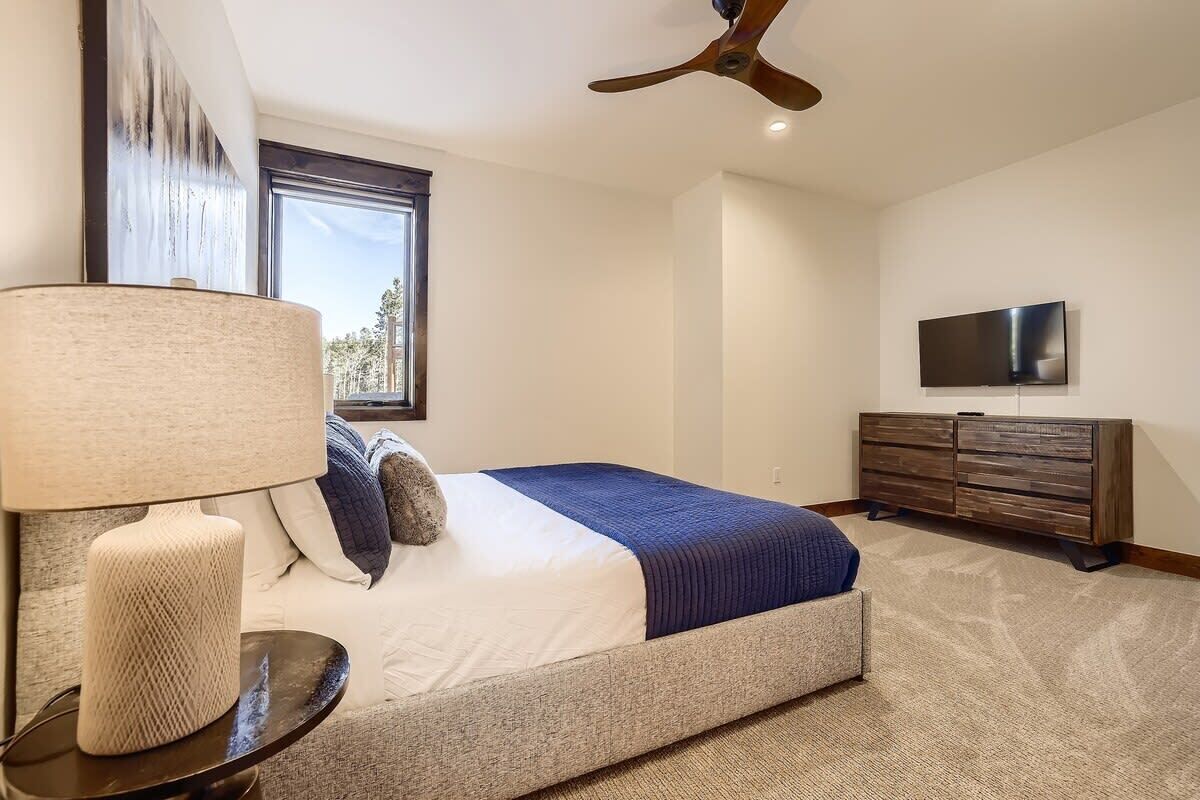 A bedroom with a bed, a TV, a ceiling fan, a window, and a lamp.