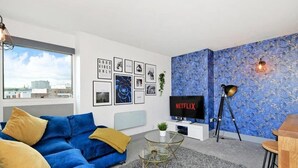 En moderne stue med en blå sofa, et sofabord i glas, et fjernsyn og en væg med billeder i rammer.