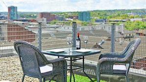 Tagterrasse med to fletstole og et bord, en flaske vin samt udsigt over bybilledet.