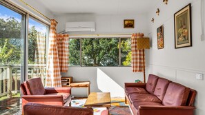 A room with a sofa, armchair, and a small table. There is a window with curtains, a wall-mounted air conditioner, and a framed picture.