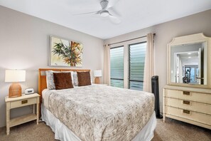 A bedroom with a bed, a nightstand, a dresser, a ceiling fan, a window with curtains, and a painting on the wall. - Oceanfront Sea Village #3212 retreat with stunning views and open living spaces (Kailua-Kona)