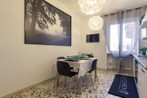 Uma sala de jantar com uma mesa posta para quatro pessoas, uma grande moldura na parede e uma área de cozinha com bancada e armários.
