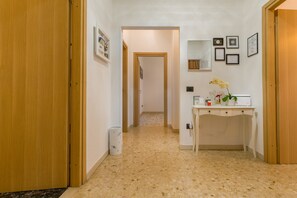 A hallway with a wooden door on the left, a desk with a vase of flowers on the right, and framed certificates on the wall.
