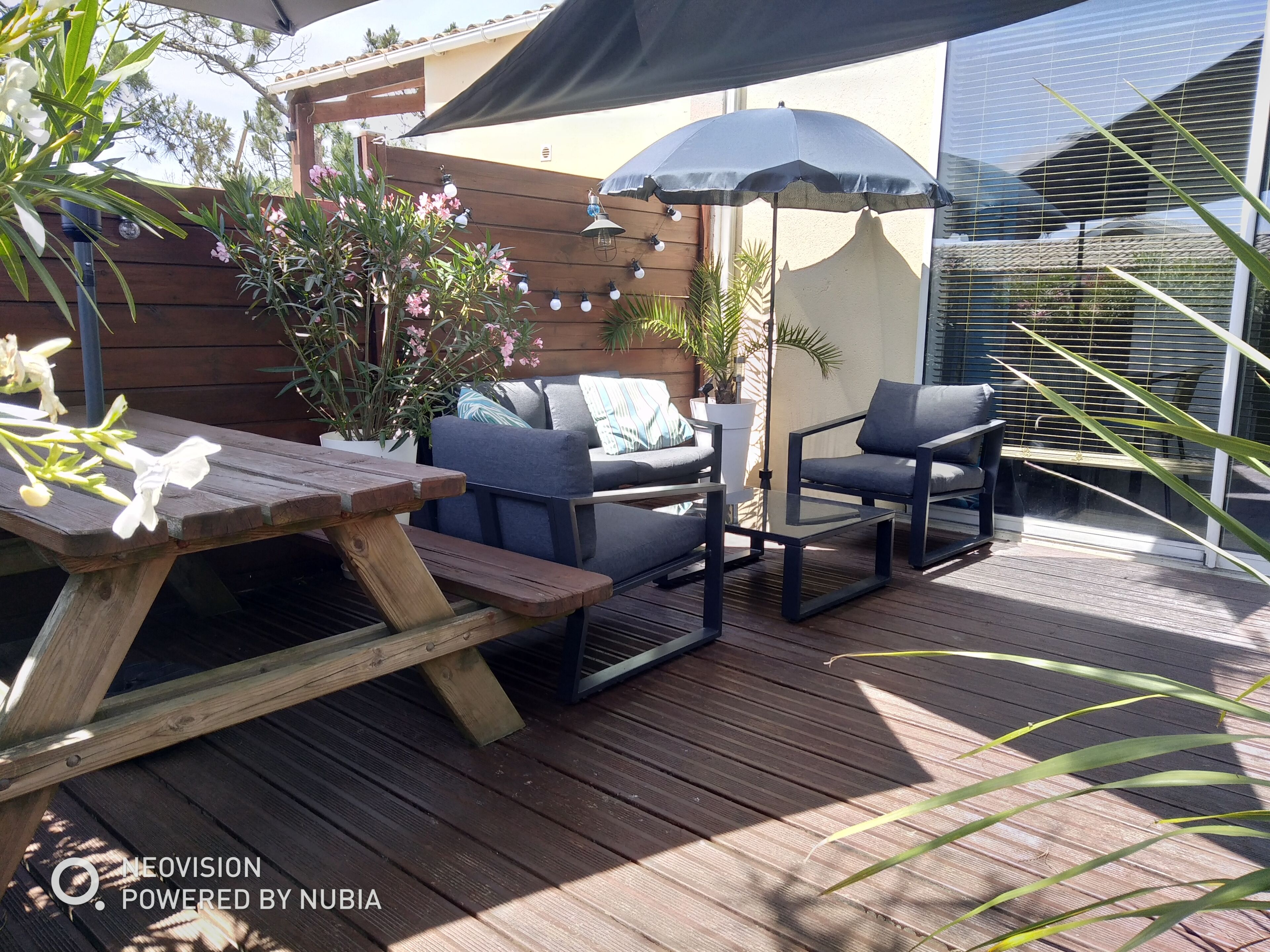 An outdoor patio with a wooden table, two chairs, a small table, and a large umbrella providing shade.