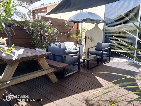 An outdoor patio with a wooden table, two chairs, a small table, and a large umbrella providing shade.