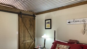 Ein Schlafzimmer mit einem Bett, Kissen, einem Nachttisch, einer Lampe, einer Holztür und einer Wand mit einem gerahmten Zitat und einem Schild.
