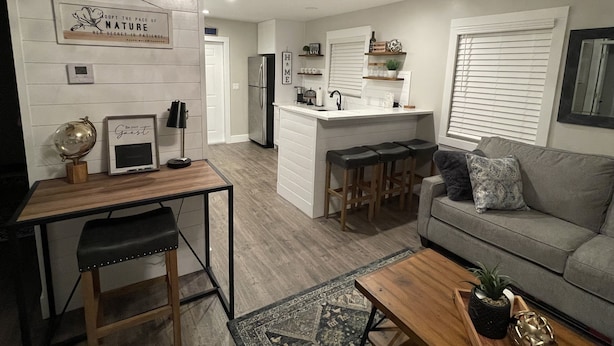 A modern living room with a grey sofa, a coffee table, and a small bar area with stools. There is a kitchen in the background with a refrigerator and a sink.