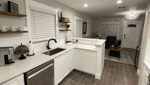 A modern kitchen with white cabinetry, a dishwasher, and a built-in oven. There is a window with blinds, a countertop with a coffee machine, and a small plant on a shelf.