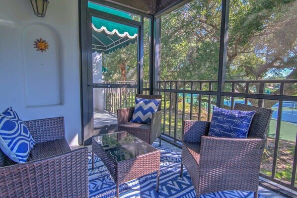 Relax in a serene outdoor space with wicker furniture and a glass railing offering views of lush greenery and a nearby tennis court.