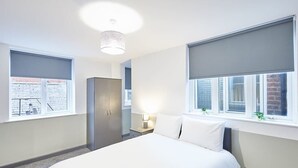 Ein modernes Hotelzimmer mit einem großen Bett, einem Kleiderschrank, einem Nachttisch mit Lampe und Fenstern mit Jalousien.