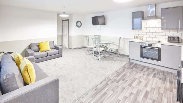 Ein modernes Wohnzimmer mit grauem Sofa, einem Esstisch mit Stühlen sowie einer Küchenzeile inklusive Fernseher, Backofen und Mikrowelle.