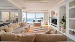 Un salon moderne avec un grand canapé beige, un tapis coloré et un téléviseur posé sur un meuble. On y trouve également un coin repas avec une table et des chaises, ainsi qu’une grande fenêtre offrant une vue sur la mer.