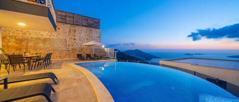A infinity pool with a view of the sea and mountains at dusk.