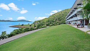A hotel with a view of a lake and mountains, a grassy lawn, and a pathway leading to the water. - Frangipani Lodge 203, Frangipani Lodge 203 (Hamilton Island)