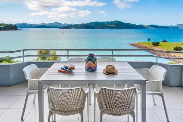 A terrace with a white table and wicker chairs, overlooking a lake with mountains in the background.