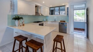 A modern kitchen with white cabinetry, a large island with stools, a stainless steel range hood, and a view to the outside. - Frangipani Lodge 203, Frangipani Lodge 203 (Hamilton Island)
