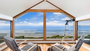 Una habitación con un amplio ventanal panorámico que da al océano, un telescopio sobre trípode y dos sillones.