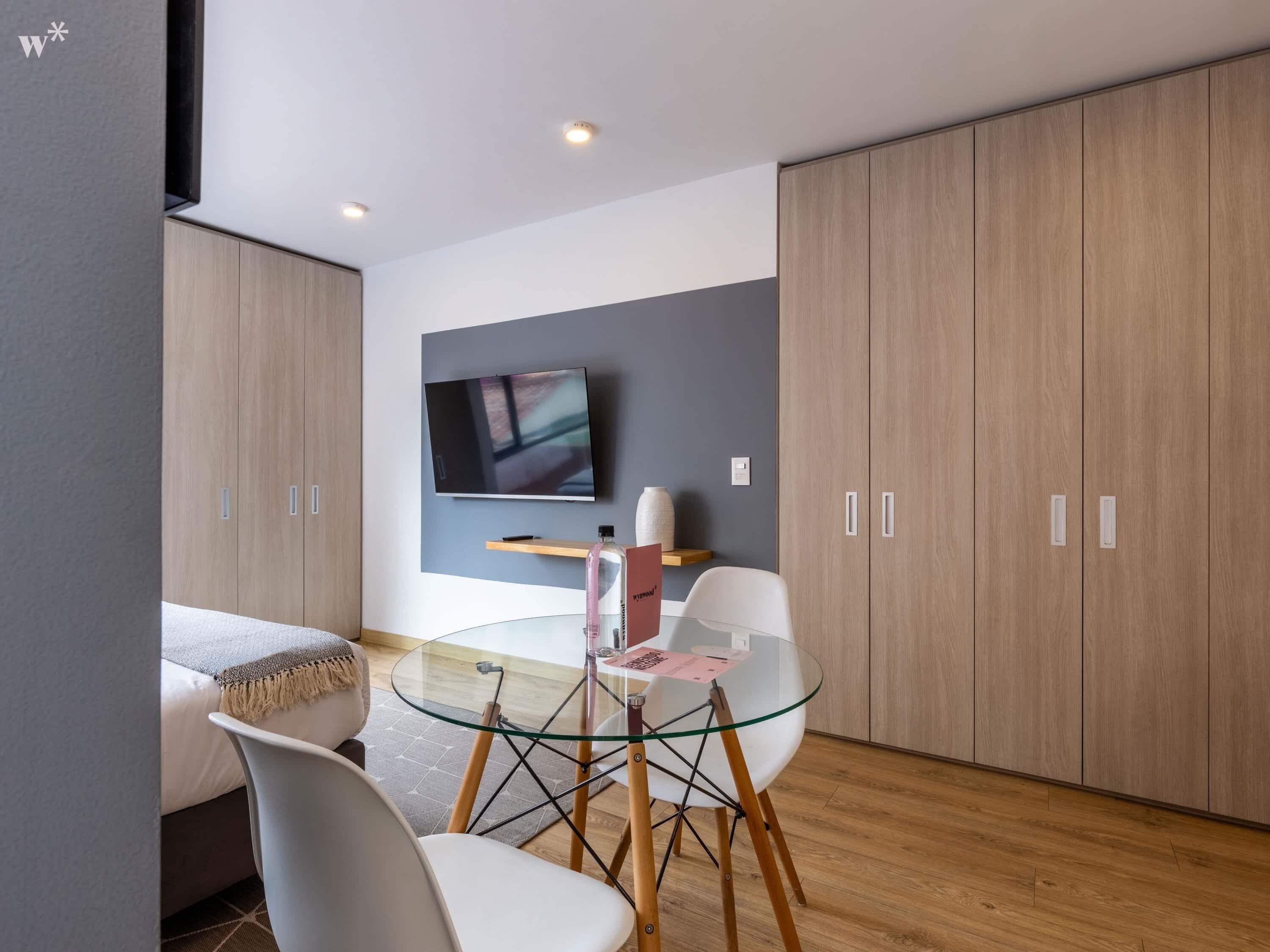 Una habitación de hotel moderna con una mesa de comedor de vidrio, sillas blancas, un televisor de pantalla plana montado en la pared y armarios de madera.