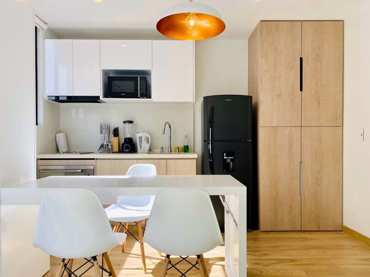 Een moderne keuken met witte kasten, een zwarte koelkast en een houten kastje. Er is een eethoek met twee witte stoelen en een tafel.