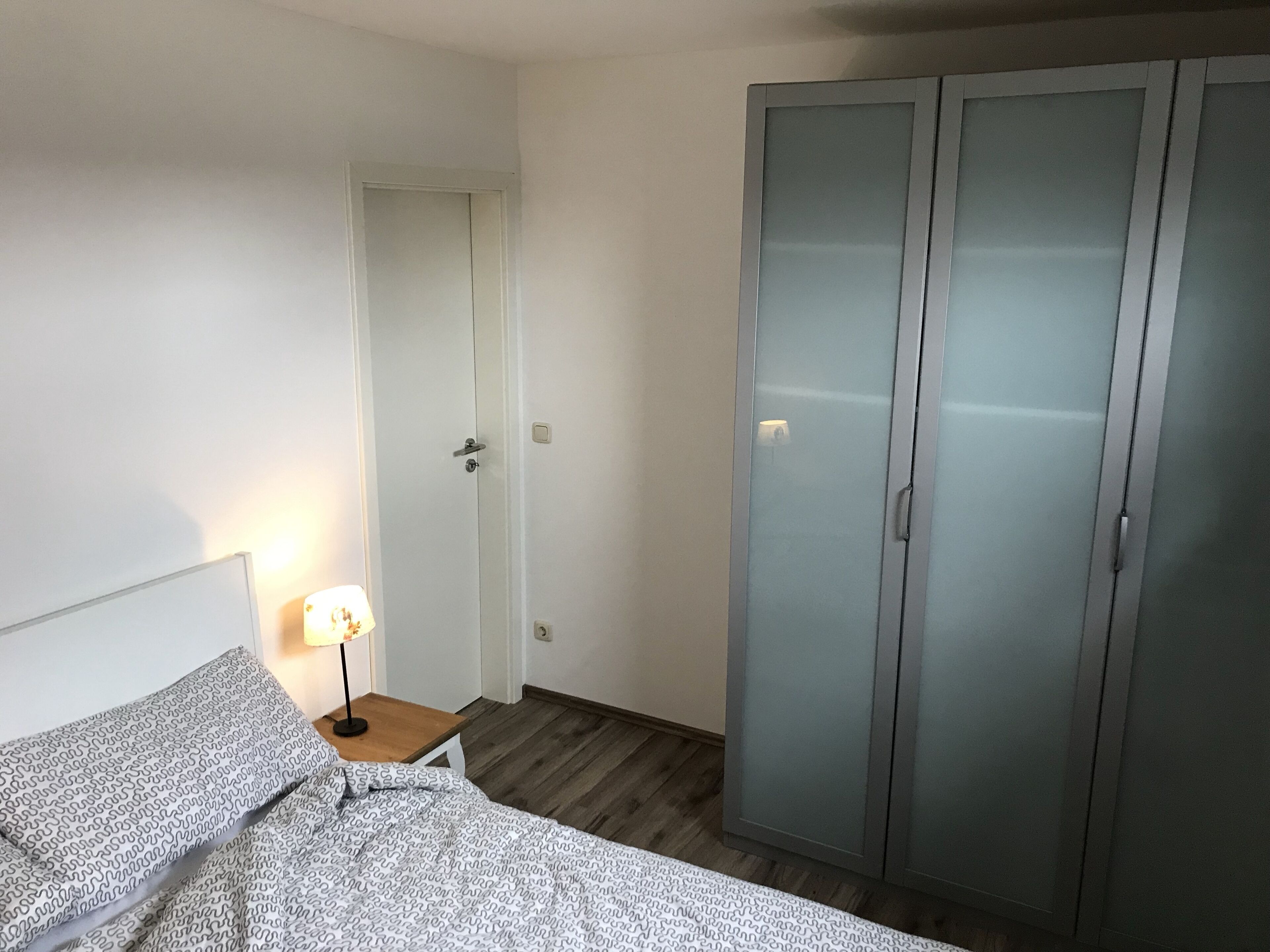 Een slaapkamer met een bed, een nachtkastje met een lamp, een kledingkast en een deur.