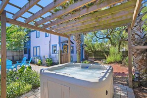 Ein Whirlpool unter einer Holzpergola, im Hintergrund ein blaues Haus und eine Terrassenfläche mit Stühlen.