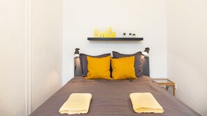 A neatly made bed with two pillows, a bedside table with a lamp, and a shelf with a 'Smile' sign above it.