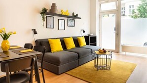 Una sala de estar moderna con un sofá gris, cojines amarillos, una mesa de comedor con sillas, una mesita auxiliar con alfombra amarilla y un ventanal con vistas al exterior.