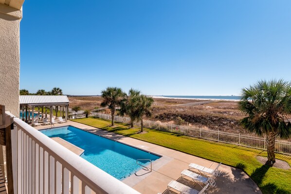 Piscina con sillas de descanso y un pabellón, rodeada por una zona vallada con palmeras y vistas a la playa.