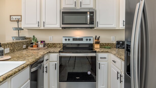 A kitchen with white cabinets, a stainless steel refrigerator, a microwave, an oven, and a granite countertop. - Gorgeous Beach Club Condo! Spectacular Views! - RAMBL ONE (Dauphin Island)