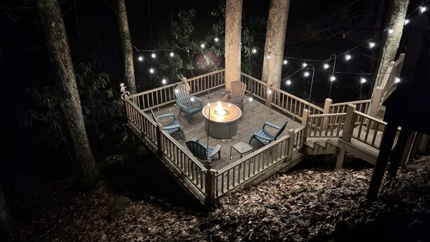 Una terraza de madera con un pozo de fuego, rodeada de sillas y luces colgantes, ubicada en un área boscosa.
