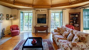 A spacious living room with a wooden ceiling, a stone fireplace, a television, a red armchair, a floral sofa, and a bookshelf.
