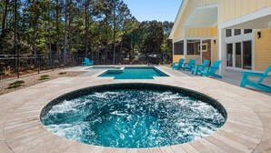 A swimming pool with a hot tub, surrounded by a deck with chairs, and a building with large windows. - Breeze The Day: Saltwater Pool, Pool Table, Spa (Corolla)