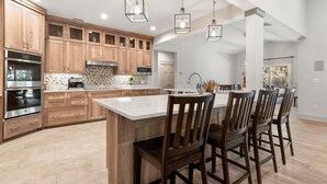 A spacious kitchen with wooden cabinets, stainless steel appliances, and a large island with bar stools. - Breeze The Day: Saltwater Pool, Pool Table, Spa (Corolla)