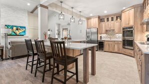 A modern kitchen with wooden cabinets, a central island, and a dining area with chairs. - Breeze The Day: Saltwater Pool, Pool Table, Spa (Corolla)