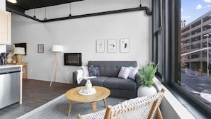 Ein modernes Wohnzimmer mit einem grauen Sofa, einem Holztisch und einem Korbstuhl. Auf dem Tisch steht eine weiße Vase, ein Stehschrank und ein Flachbildfernseher an der Wand.