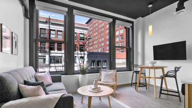 A modern living room with a sofa, a round coffee table, and a chair. There is a large window with a view of buildings outside. - Urban Modern Living Downtown w/ Free Coffee & WiFi (Spokane)