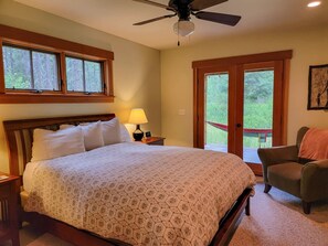 A bedroom with a bed, a nightstand, a lamp, a chair, a ceiling fan, and a view through a door.