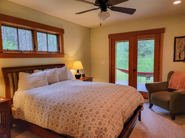 Une chambre à coucher comprenant un lit, une table de chevet, une lampe, une chaise, un ventilateur de plafond et une vue sur l’extérieur, visible à travers une porte.