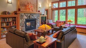 A cozy living room with a stone fireplace, leather sofas, a bookshelf, and large windows offering a view of the outdoors. - Triple J Cabin (Mazama)
