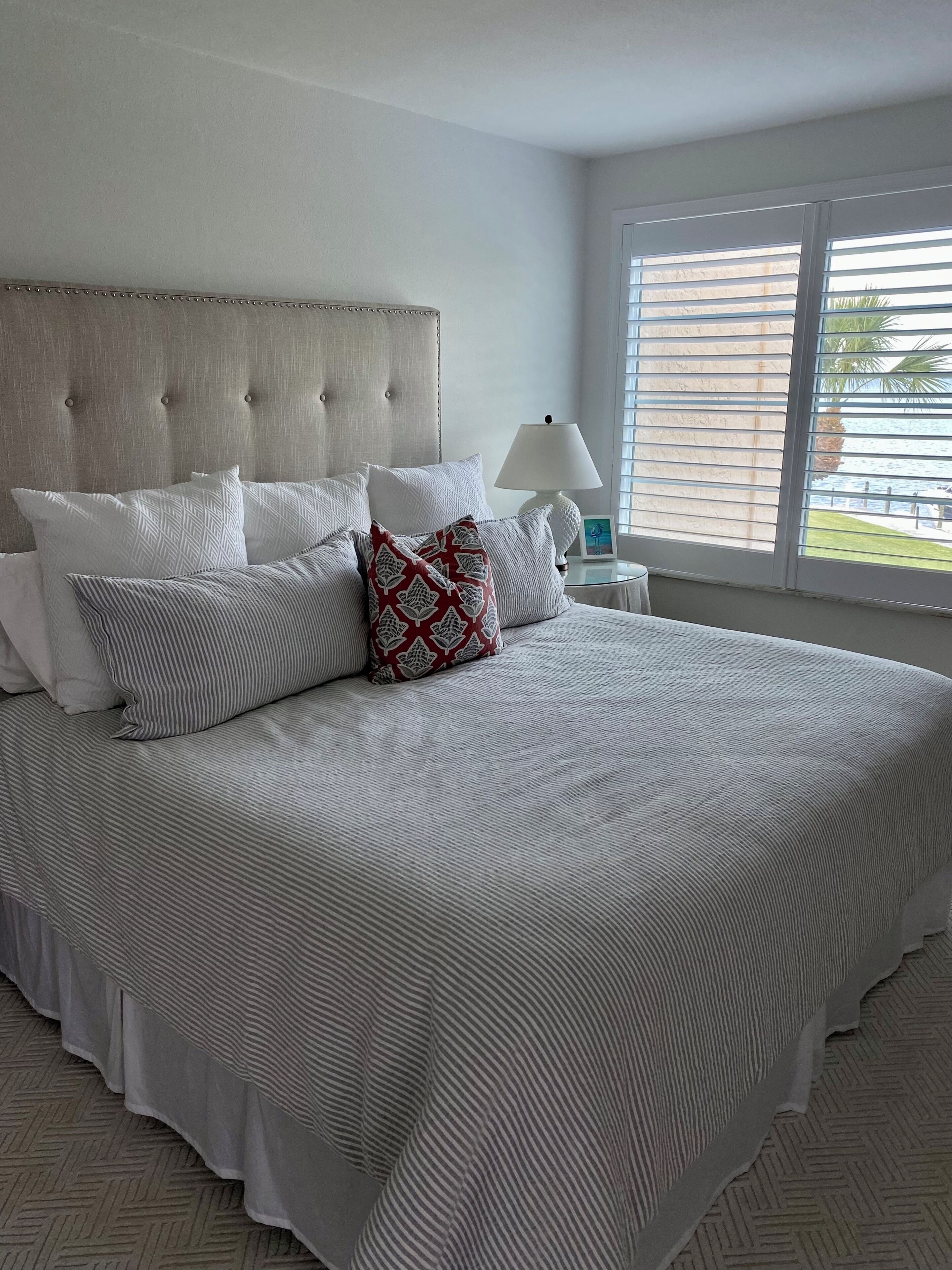 A neatly made bed with a striped bedspread and multiple pillows, a bedside lamp, and a window with blinds.