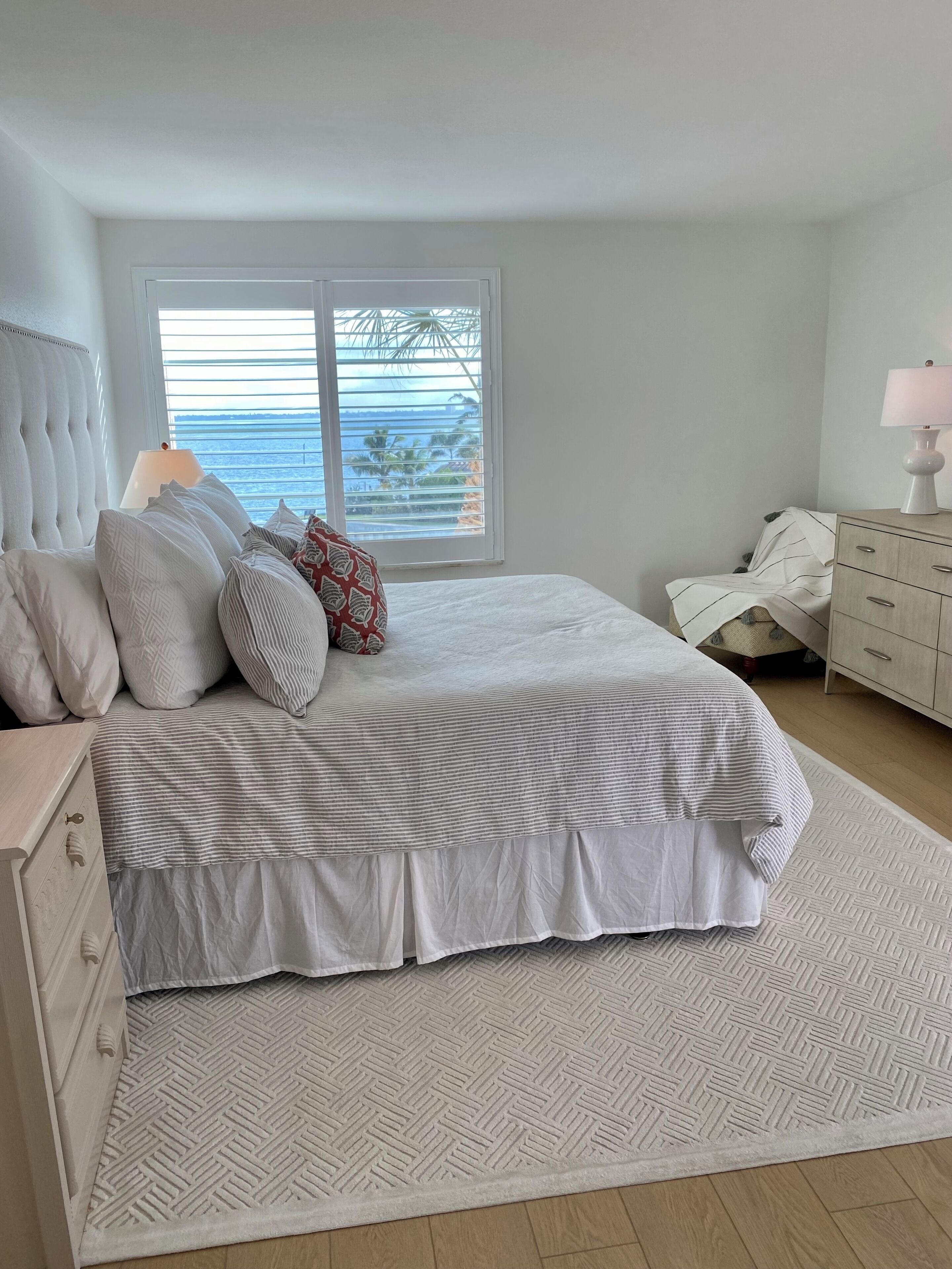 A bedroom with a large bed, a nightstand, a dresser, a window with blinds, and a view of the outdoors.