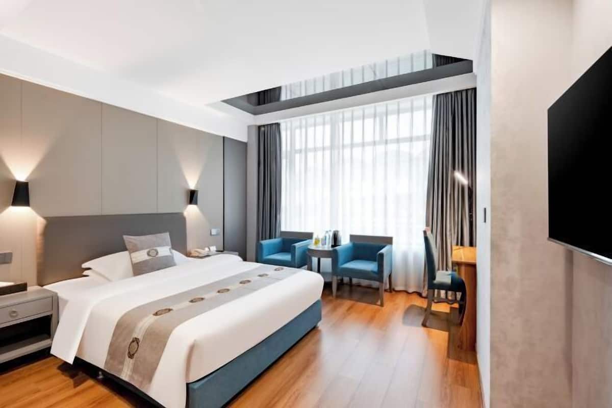 A modern hotel room with a large bed, a desk, two chairs, a TV, and a window with curtains.